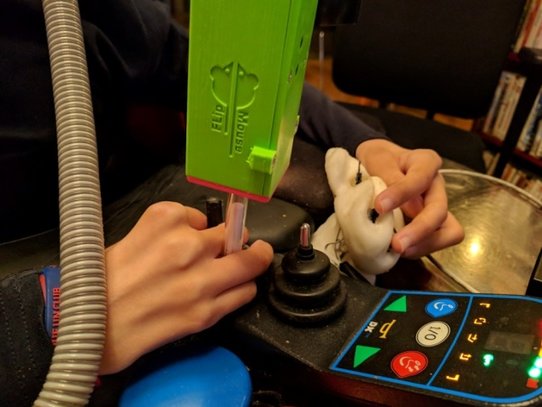 Bild zeigt die Verwendung der FlipMouse als Fingerjoystick. Das Gerät ist so montiert, dass das Mundstück-Röhrchen die Hand berührt und durch kleine Bewegungen ausgelenkt werden kann.